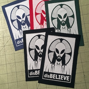 Può includere: Quattro toppe termoadesive in bianco e nero con una grafica di un alieno con un'aureola e il testo "disBELIEVE". Le toppe sono su una superficie a scacchi blu e bianca.