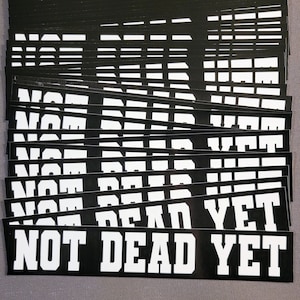 May include: A stack of black and white stickers with the text "NOT DEAD YET" in white letters. The stickers are arranged in a staggered pattern.