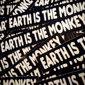 Könnte beinhalten: Nahaufnahme eines Stapels schwarzer und weißer Aufkleber mit dem Text "Earth is the monkey", der auf jedem Aufkleber wiederholt wird.