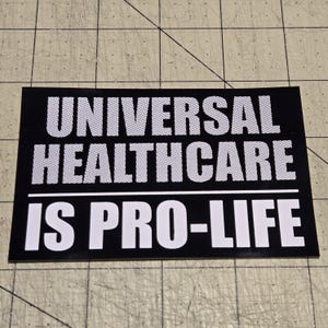 Puede incluir: Una pegatina rectangular negra con texto blanco. El texto dice "UNIVERSAL HEALTHCARE" encima de "IS PRO-LIFE". Las palabras están en negrita, con una fuente sans-serif. La pegatina está sobre una superficie gris claro con un patrón de cuadrícula.