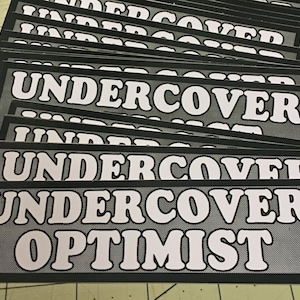 Op de afbeelding: Een stapel zwart-wit stickers met de tekst "UNDER COVER OPTIMIST" in wit op een zwarte achtergrond.