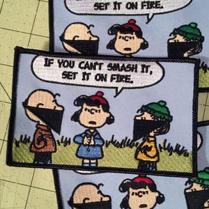 May include: A cartoon patch featuring a speech bubble that reads "If you can't smash it, set it on fire." The patch depicts three characters from the Peanuts comic strip: Charlie Brown, Lucy, and Linus. Charlie Brown is wearing a brown jacket and a black bandana. Lucy is wearing a blue dress and a red beret. Linus is wearing a yellow jacket and a green hat. The patch is embroidered on a blue background.