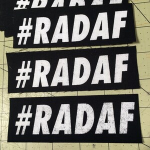 Puede incluir: Tres parches de tela negra con texto blanco que dice "#RADAF" en una fuente en negrita y en bloque. Los parches están cosidos juntos y tienen un aspecto ligeramente desgastado.