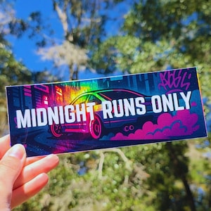 May include: A rectangular sticker with the text "MIDNIGHT RUNS ONLY" in white, featuring a stylized car in a vibrant, colorful design. The car is depicted in shades of pink, green, and blue, with a cityscape background and graffiti art.
