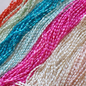 May include: A close-up of several strands of colorful beads. The beads are in shades of orange, light green, turquoise, pink, and white. The beads are oval-shaped and appear to be strung together. The background is a book page.