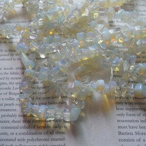 May include: A necklace made of white and translucent opal gemstone beads. The beads are irregular in shape and size, and some have a slight yellow hue. The necklace is lying on a page of a book with text about Renaissance necklaces.