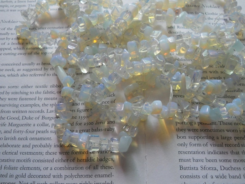 May include: A necklace made of white and translucent opal gemstone beads. The beads are irregular in shape and size, and some have a slight yellow hue. The necklace is lying on a page of a book with text about Renaissance necklaces.