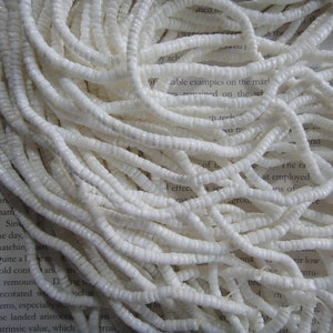 Puede incluir: Una hebra de cuentas de concha blanca, cada cuenta es una forma ovalada plana y pequeña con una superficie ligeramente texturizada.