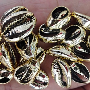 May include: A collection of gold-colored cowrie shells, a type of sea shell, with a shiny finish. The shells are arranged in a pile, showing their intricate details.