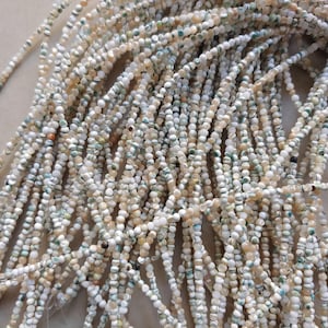 Puede incluir: Primer plano de múltiples hebras de pequeñas cuentas redondas en tonos blanco, beige y verde pálido. Las cuentas están ensartadas, creando una apariencia texturizada. Las cuentas probablemente están hechas de materiales naturales, como conchas o piedras.