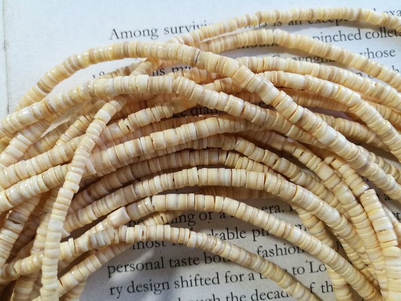 May include: A close-up of a strand of cream-colored shell beads. The beads are small and round, and they are arranged in a loose pile.