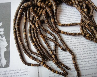 May include: Close-up of a necklace made of small, round, dark brown and tan wooden beads. The beads are strung together, creating multiple strands. The necklace is displayed on a printed page with text and a black and white image.