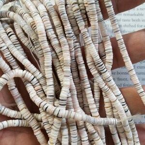 May include: A strand of white, brown, and black shell beads. The beads are round and flat, and they are strung on a white thread.