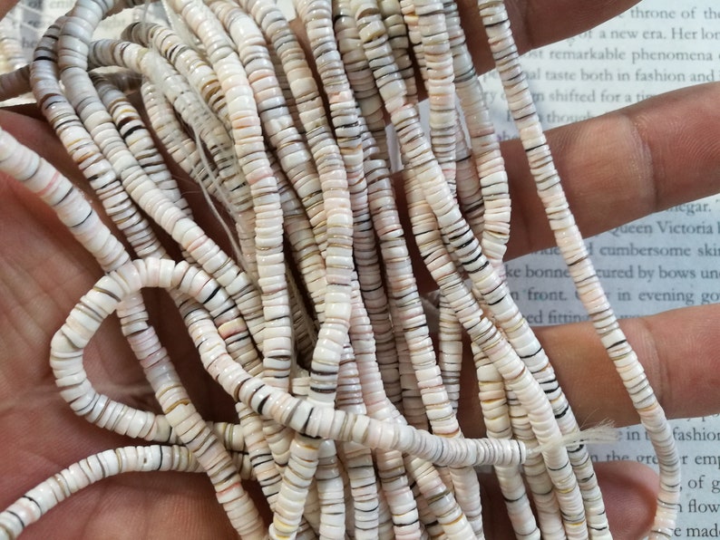 May include: A strand of white, brown, and black shell beads. The beads are round and flat, and they are strung on a white thread.