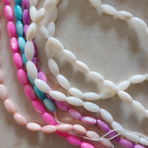 May include: A close-up of five strands of oval-shaped beads. The beads are made of white, pink, light blue, and purple iridescent shell. The beads are strung on a white thread.