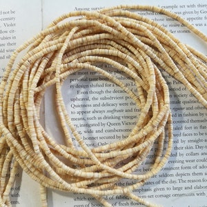 May include: A close-up of a strand of cream-colored shell beads. The beads are small and round, and they are arranged in a long strand. The beads are lying on a book with text about fashion and design.