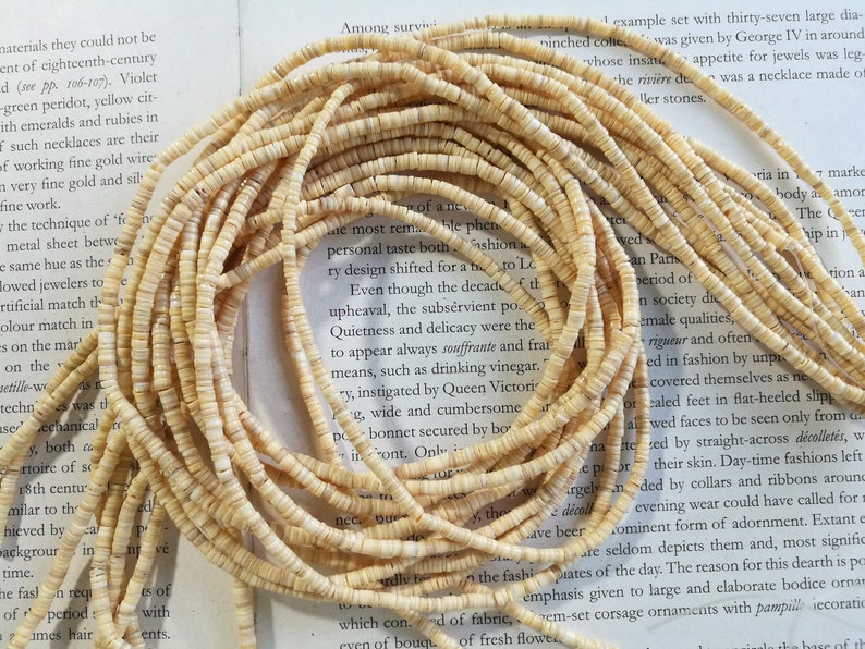 May include: A close-up of a strand of cream-colored shell beads. The beads are small and round, and they are arranged in a long strand. The beads are lying on a book with text about fashion and design.
