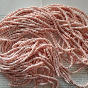 May include: A pile of pink and white disc-shaped beads, likely for jewelry making, are arranged in a tangled bundle. The beads are strung together, and the ends are tied with a clear string. The background is a page of printed text.