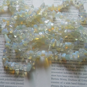 May include: A necklace made of iridescent, light blue and white opal gemstone beads. The beads are irregular in shape and size, and are strung on a clear thread. The necklace is lying on a page of a book.