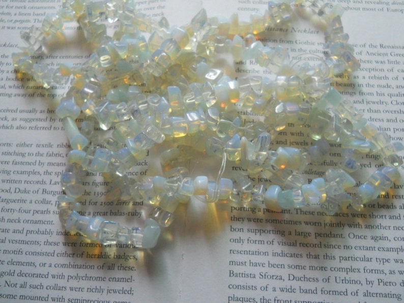 May include: A necklace made of iridescent, light blue and white opal gemstone beads. The beads are irregular in shape and size, and are strung on a clear thread. The necklace is lying on a page of a book.