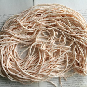 May include: A large pile of pale pink, round, and flat beads. The beads are made of a natural material, possibly shell or bone. The beads are arranged in a loose, tangled pile.