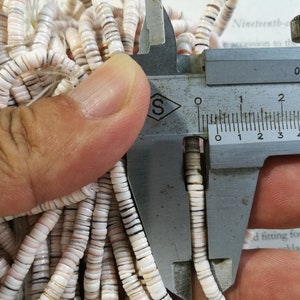 May include: Close-up of a caliper measuring the size of white and brown shell beads. The caliper reads 0.02mm.