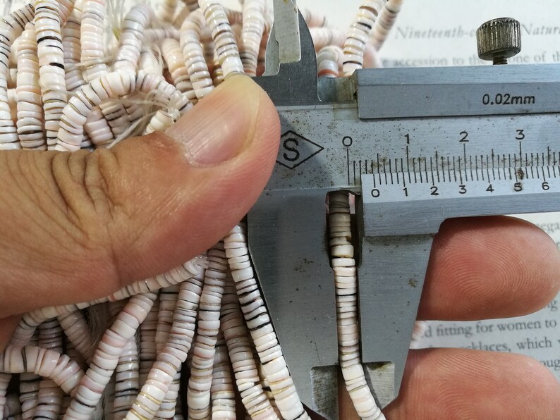 May include: Close-up of a caliper measuring the size of white and brown shell beads. The caliper reads 0.02mm.