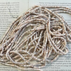 May include: A pile of white and brown shell beads. The beads are small and round, and they are arranged in a random pattern. The beads are on a white surface.