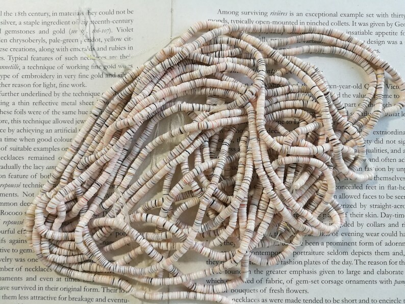May include: A pile of white and brown shell beads. The beads are small and round, and they are arranged in a random pattern. The beads are on a white surface.