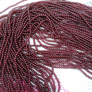 May include: A large quantity of small, round, dark red garnet beads. The beads are strung on thin threads and are arranged in a pile.