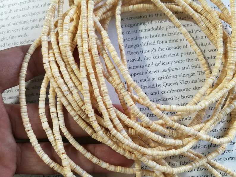 May include: A close-up of a strand of white shell beads. The beads are small and round, and they are strung together on a thin cord. The beads are arranged in a loose, flowing pattern.