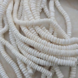Puede incluir: Una hebra de cuentas de concha blanca, hechas de pequeños trozos de concha redondos y ligeramente aplanados. Las cuentas están enhebradas en un hilo transparente, creando una hebra larga y continua.