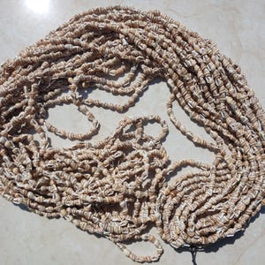 Puede incluir: Una larga hebra de cuentas de concha blancas y marrones. Las cuentas son pequeñas y de tamaño uniforme. La hebra está enrollada y enredada.