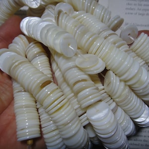 Puede incluir: Una hebra de cuentas de nácar blanco. Las cuentas son redondas y planas, con un agujero en el centro. Las cuentas están enhebradas en un hilo transparente.