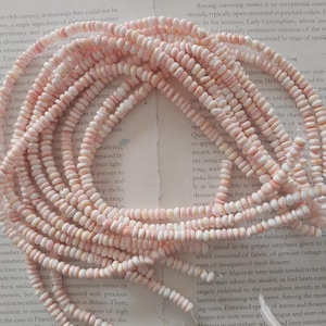 May include: A collection of pale pink beads, possibly gemstones or pearls, are arranged in a circular design. The beads are small and have an irregular shape, strung together to create a delicate look. The beads are on a background of printed text.