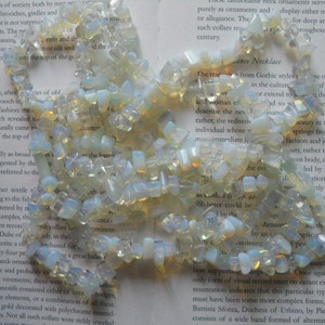 May include: A strand of iridescent white and yellow opal gemstone beads. The beads are irregular in shape and size, and they are strung together on a clear thread. The beads are scattered on a page of a book.