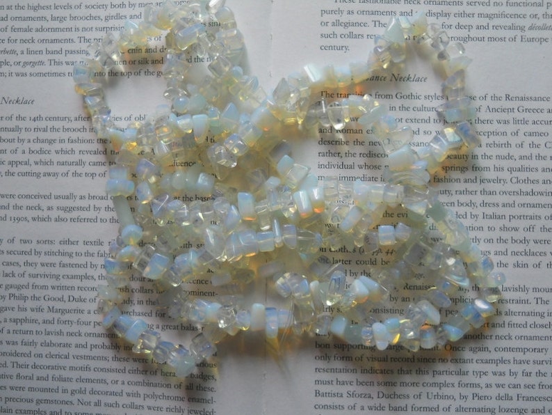 May include: A strand of iridescent white and yellow opal gemstone beads. The beads are irregular in shape and size, and they are strung together on a clear thread. The beads are scattered on a page of a book.