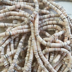 May include: A close-up of a pile of white, pink, and brown shell beads. The beads are small and round, and they have a natural, organic look.