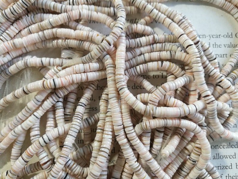 May include: A close-up of a pile of white, pink, and brown shell beads. The beads are small and round, and they have a natural, organic look.