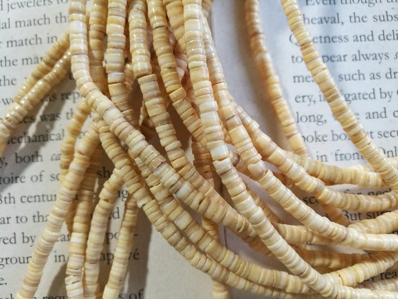 May include: A close-up of a strand of cream-colored shell beads. The beads are small and round, and they are strung together on a thin cord. The beads are arranged in a random pattern, and they are all the same size and shape.