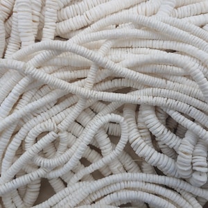 May include: A close-up of a pile of white, round, shell beads. The beads are strung together in long strands, creating a textured and organic look.
