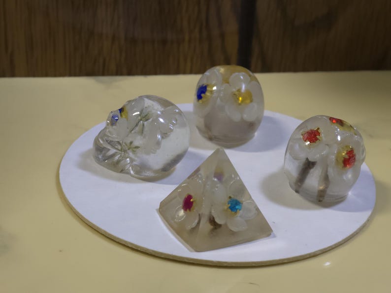 May include: Four clear resin objects featuring embedded white flowers with colorful centers. The collection includes a skull-shaped piece, two rounded dome-shaped items, and one pyramid-shaped piece, all displaying small red, blue, and yellow accents.