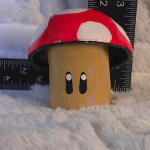 May include: A painted mushroom figurine with a red cap featuring white spots and a tan stem with two black oval eyes. A black ruler is positioned behind the object, indicating a height of approximately 2.5 inches and a width of 2.5 inches.