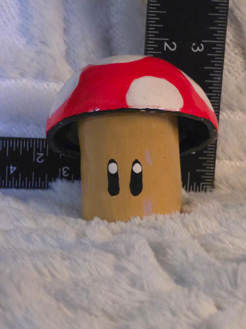 May include: A painted mushroom figurine with a red cap featuring white spots and a tan stem with two black oval eyes. A black ruler is positioned behind the object, indicating a height of approximately 2.5 inches and a width of 2.5 inches.