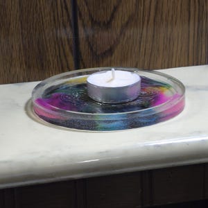 May include: A circular clear resin coaster featuring a swirling multi-colored pattern of pink, blue, and yellow hues with an embossed spiral design. A small white tea light candle with a silver metal casing sits in the center of the coaster.