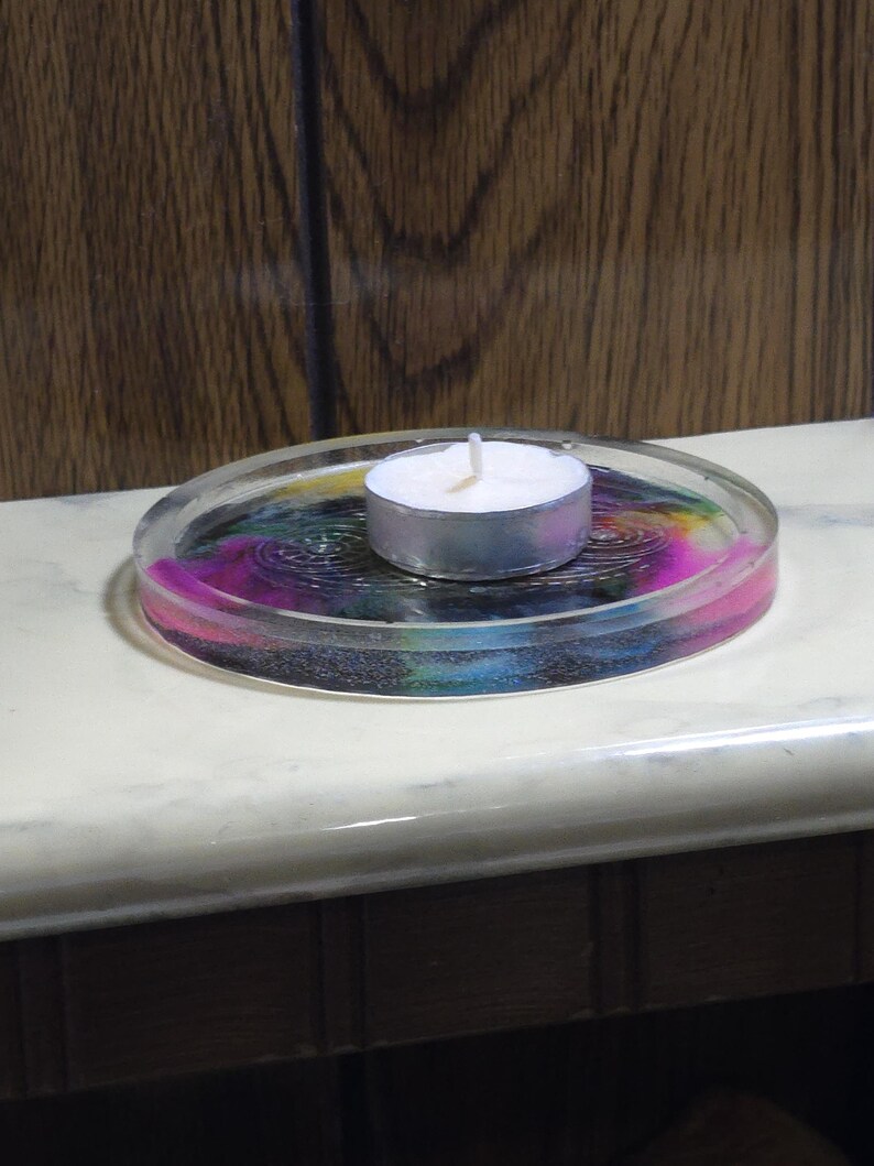 May include: A circular clear resin coaster featuring a swirling multi-colored pattern of pink, blue, and yellow hues with an embossed spiral design. A small white tea light candle with a silver metal casing sits in the center of the coaster.