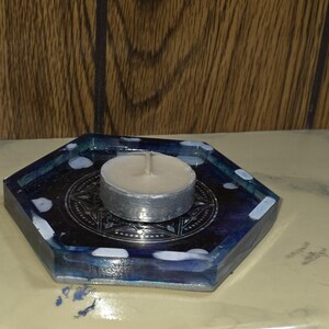 May include: A hexagonal resin tray featuring a dark blue and black marbled pattern with white accents. A silver-colored metal tea light candle holder with a geometric star design sits in the center, holding a small white wax tea light candle.
