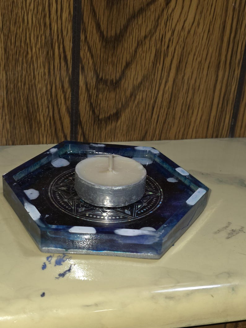 May include: A hexagonal resin tray featuring a dark blue and black marbled pattern with white accents. A silver-colored metal tea light candle holder with a geometric star design sits in the center, holding a small white wax tea light candle.