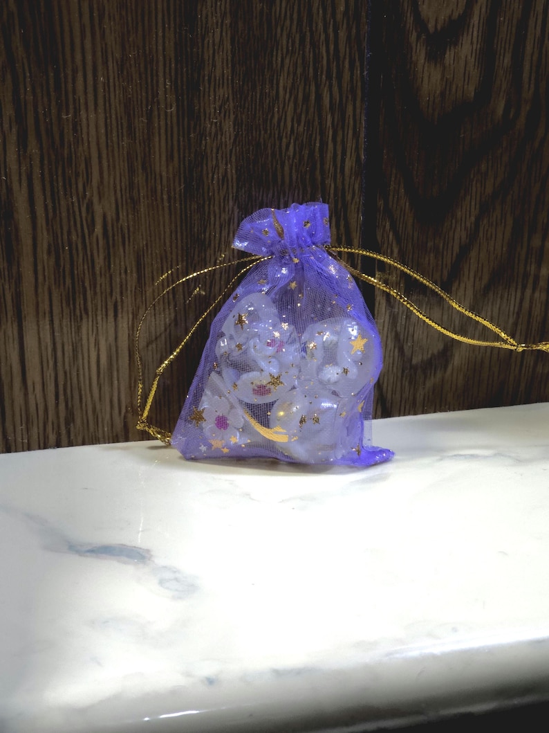 May include: A small purple organza drawstring pouch featuring a gold star pattern. The sheer fabric reveals several white, rounded, opaque objects inside. The bag is cinched at the top with thin gold cords, creating a gathered effect at the opening.