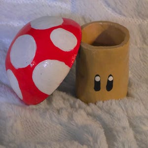 May include: A cylindrical tan container with two black and white oval eyes painted on the front, accompanied by a separate red mushroom cap lid featuring white circular spots. The object is constructed with a hollow interior and a smooth finish.